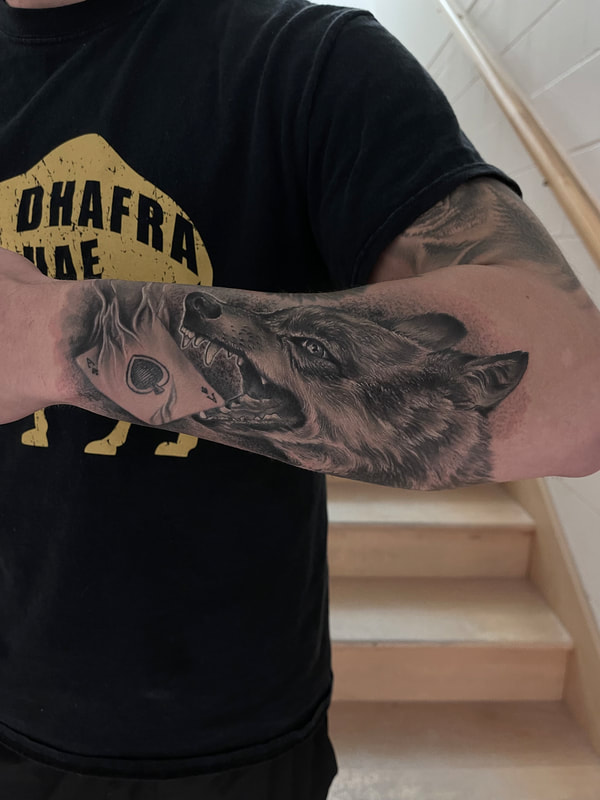 Realistic black and grey tattoo of a wolf biting an ace card on a forearm.